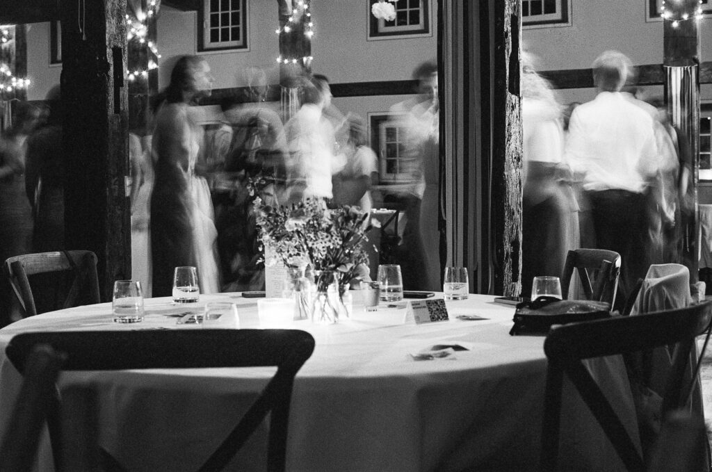 Analoge Schwarz-weiß-Fotografie einer Hochzeitsfeier mit starker Filmkörnung; im scharfen Vordergrund steht ein gedeckter Tisch, während die tanzenden Gäste im Hintergrund in ästhetischer Bewegungsunschärfe verschwimmen.